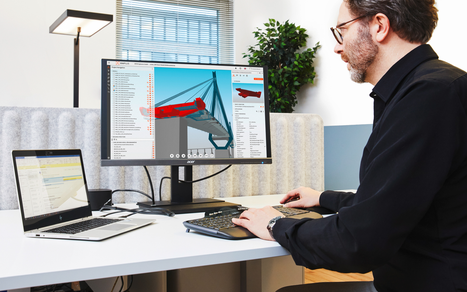 Person sitzt an einem Schreibtisch mit zwei Bildschirmen, auf dem großen Monitor ist ein 3D-BIM-Modell einer Brücke zu sehen.