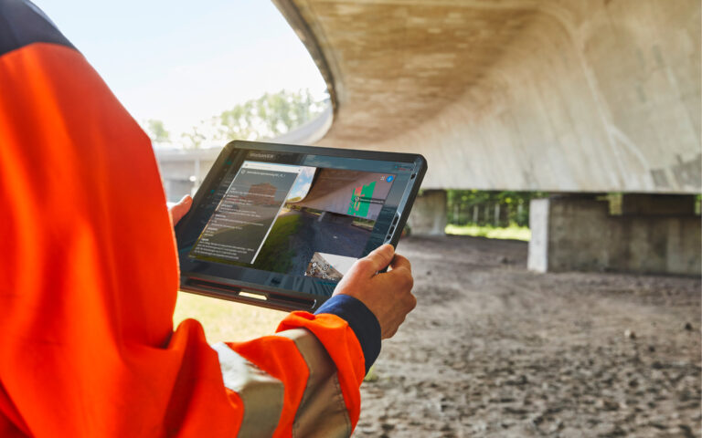 Person in orangefarbener Warnschutzjacke hält ein Tablet mit einer technischen 3D-Visualisierung unter einer Brücke.