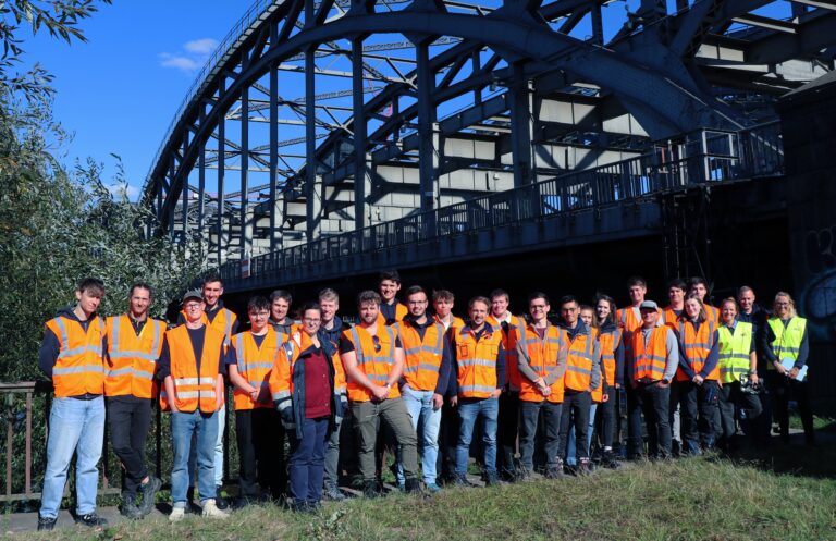 Gruppe von Personen in Warnwesten steht vor einer großen Stahlbogenbrücke im Freien.