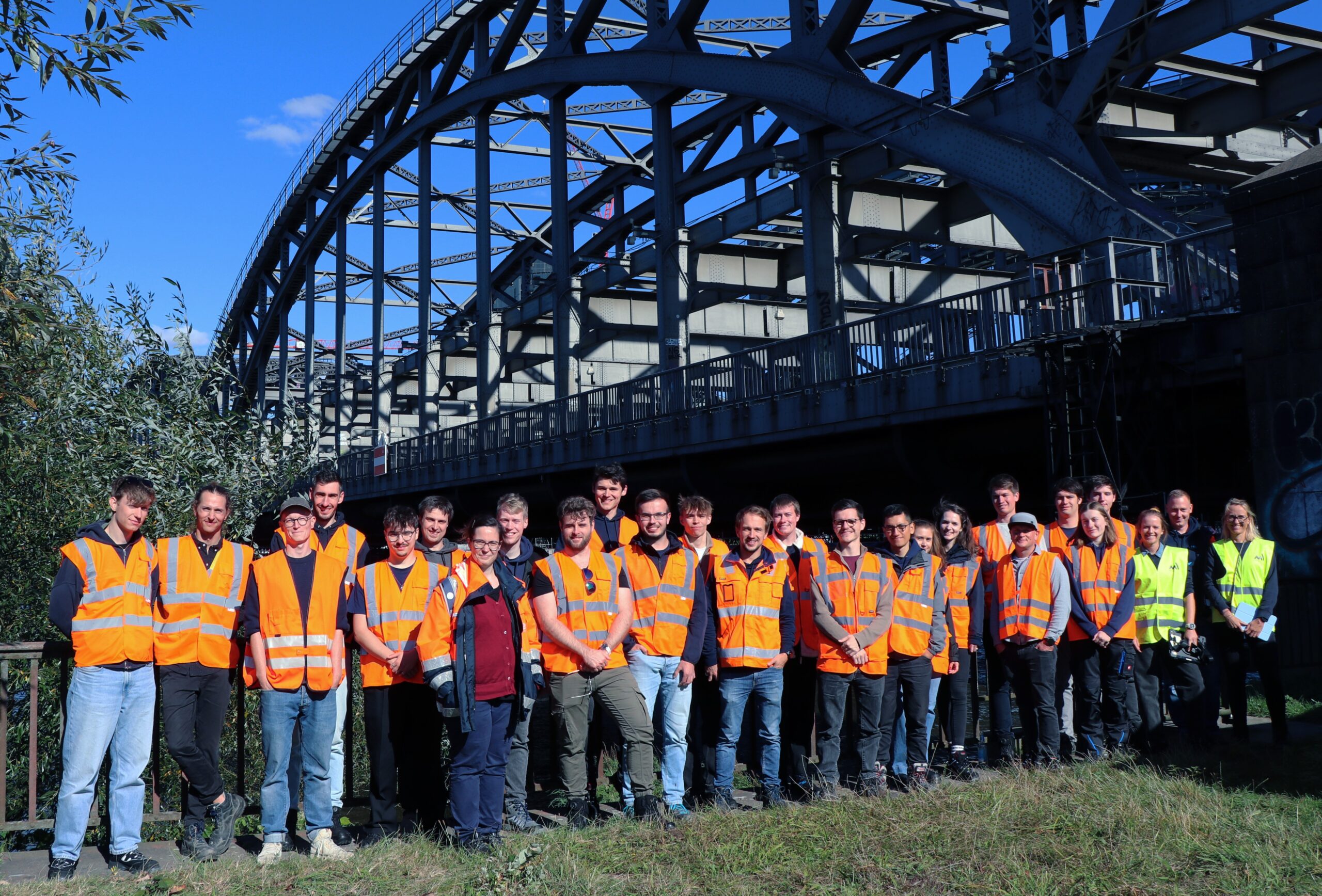 Gruppe von Personen in Warnwesten steht vor einer großen Stahlbogenbrücke im Freien.