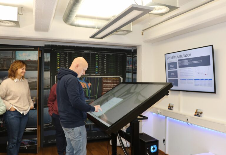 Mehrere Personen stehen in einem Raum, eine Person bedient einen großen, waagerecht geneigten Touchscreen, an der Wand ist ein Bildschirm mit einer Verkehrssimulation zu sehen.