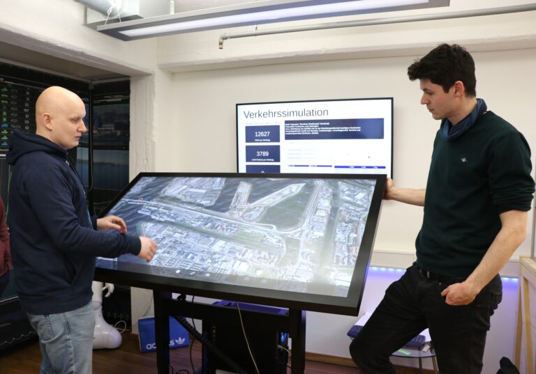 Zwei Männer stehen an einem großen Touchscreen-Tisch mit einer Luftaufnahme einer Verkehrssituation, im Hintergrund ein Bildschirm mit der Überschrift 'Verkehrssimulation'.