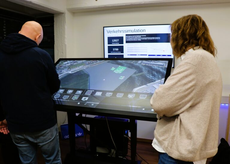 Zwei Personen stehen vor einem großen Touchscreen mit einer Verkehrssimulation, im Hintergrund ein Bildschirm mit Diagrammen und Zahlen.