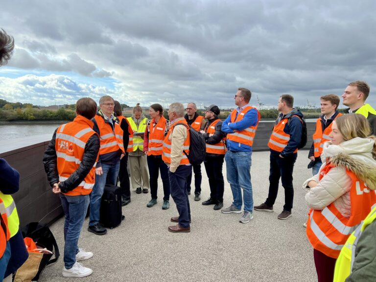 Gruppe von Menschen mit orangen Warnwesten steht auf einer Aussichtsplattform am Wasser unter bewölktem Himmel.