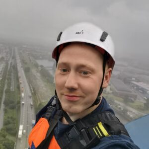 Person mit weißem Schutzhelm und Sicherheitsgeschirr auf einer hohen Plattform mit Blick auf eine Straße und Industriegebäude im Nebel.