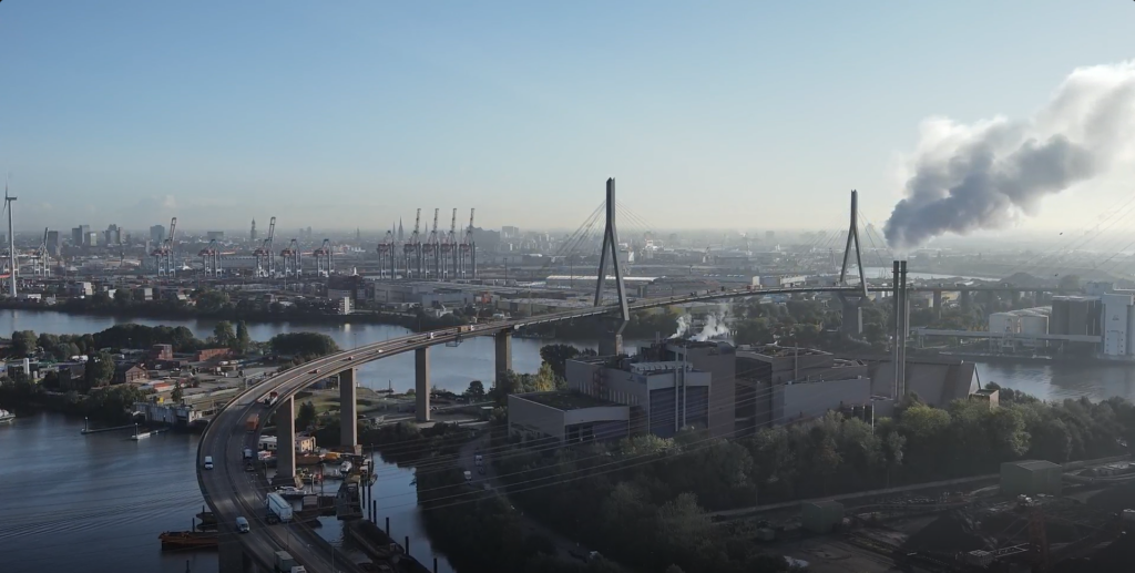 Blick auf eine Hafenanlage mit einer großen Brücke, die über Wasser führt, und einem Industriegebäude, aus dessen Schornstein Rauch aufsteigt